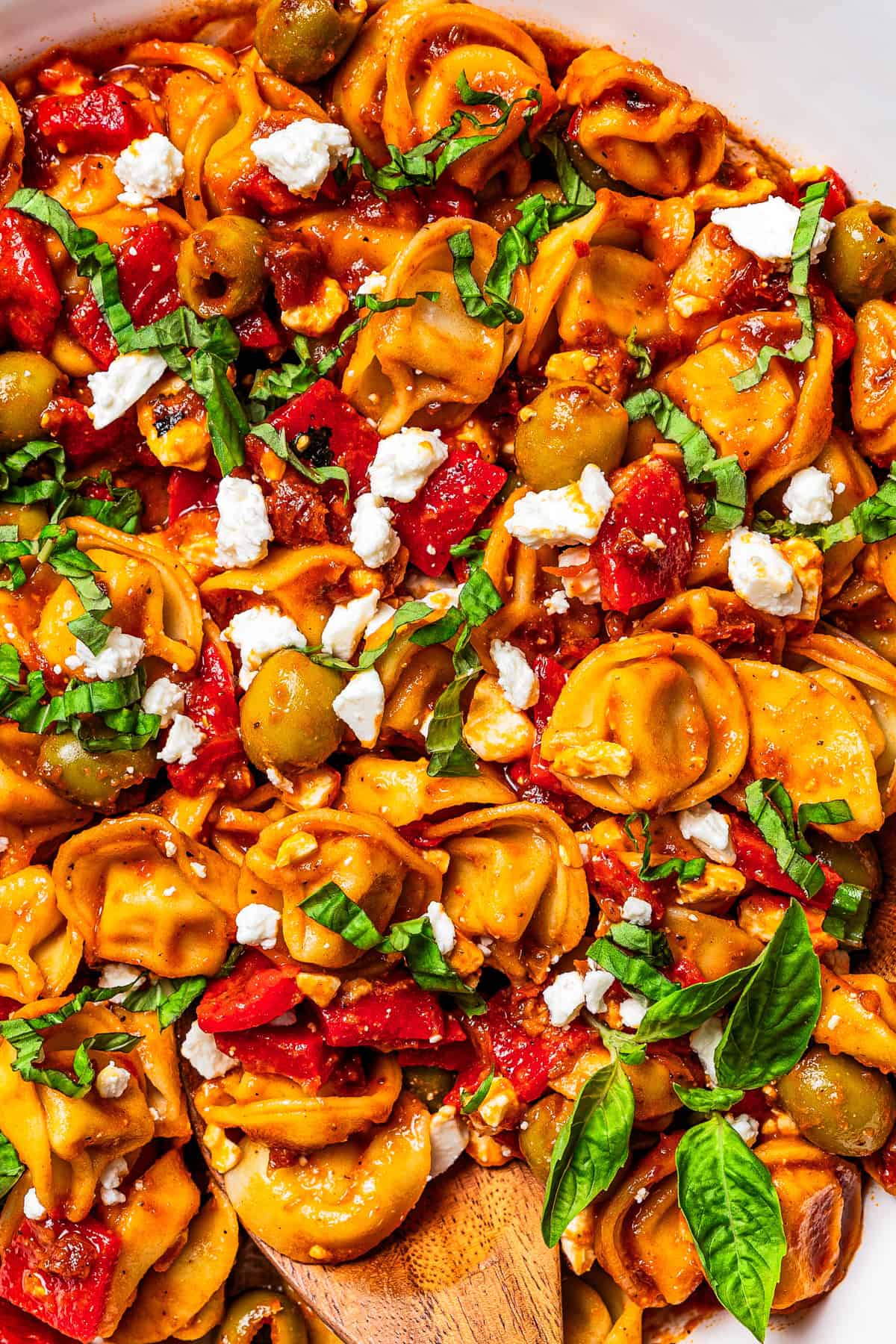 This Mediterranean tortellini pasta salad is a flavorful, easy summer side dish with cheesy tortellini, olives, red peppers, and feta cheese! Close-up of cooked tortellini salad tossed with tomatoes, olives, crumbled cheese, and fresh basil.