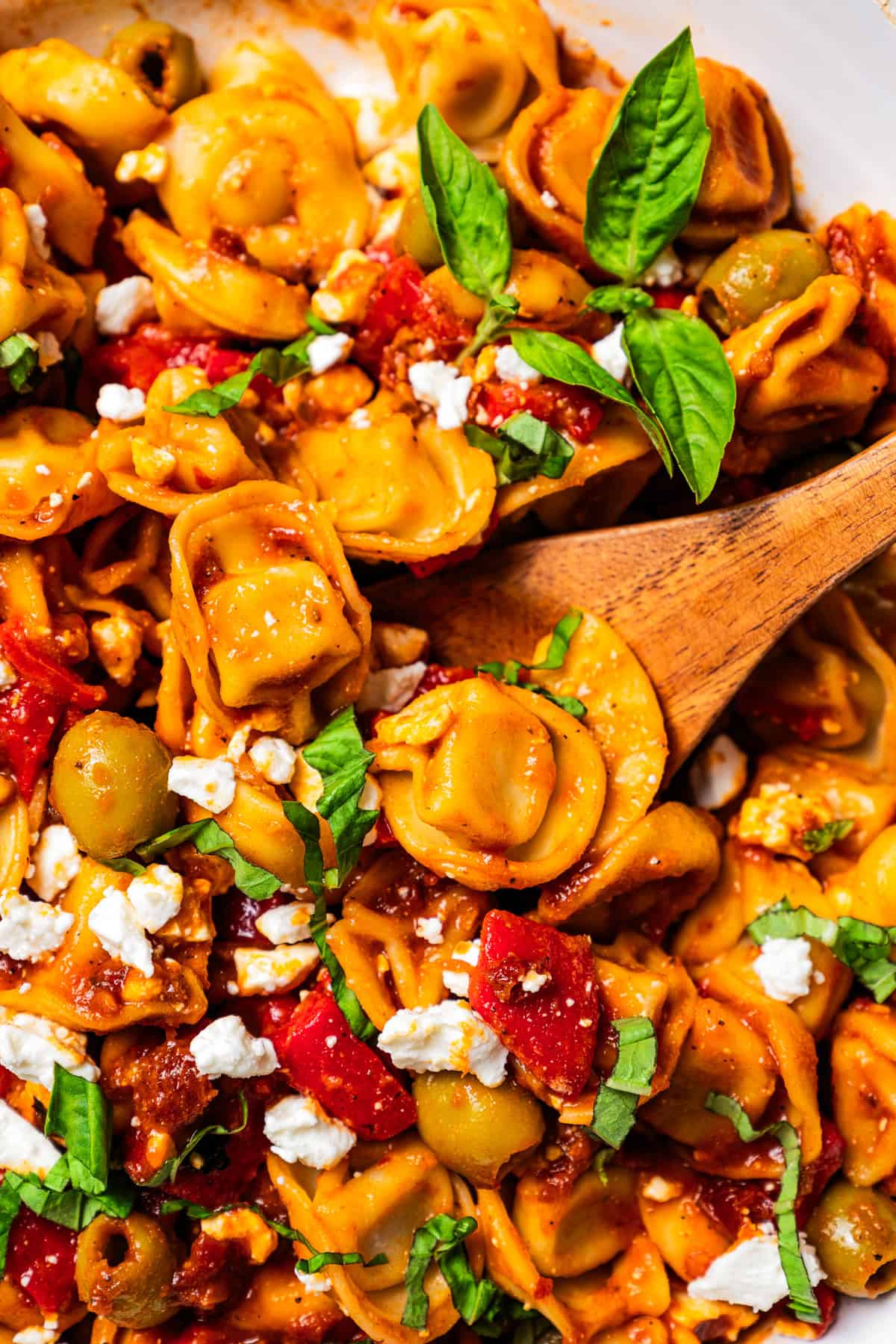 This Mediterranean tortellini pasta salad is a flavorful, easy summer side dish with cheesy tortellini, olives, red peppers, and feta cheese! Wooden spoon stirring through a large bowl of tortellini pasta salad.