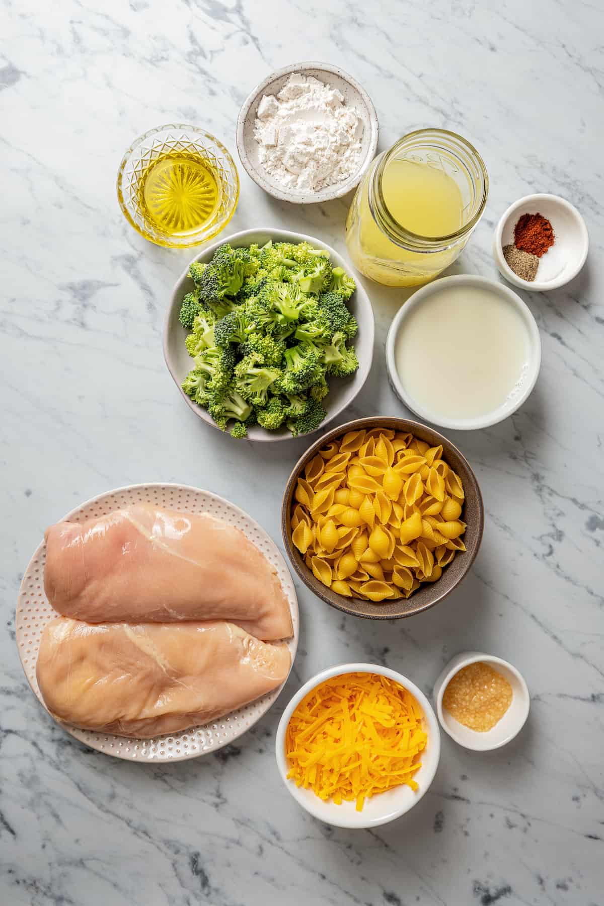 Overhead view of the ingredients needed for chicken broccoli pasta: a plate with raw chicken breasts, a bowl of shredded cheddar cheese, a bowl of broccoli, a bowl of pasta shells, a bowl of milk, a jar of chicken broth, a bowl of olive oil, a bowl of flour, a bowl of garlic, and a bowl of salt, pepper, and chili powder