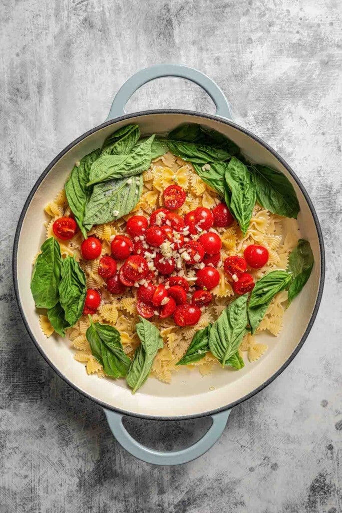 Halved cherry tomatoes, basil, and garlic added to a pot with pasta and water.