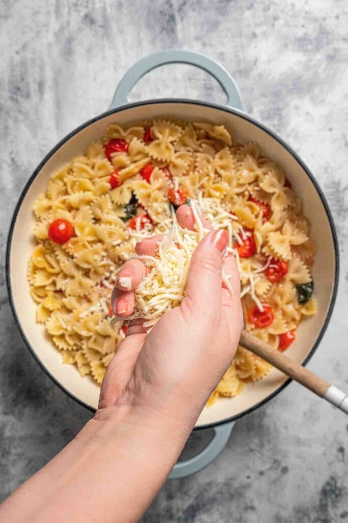 A hand sprinkling shredded mozzarella cheese into a pot of cooked pasta.