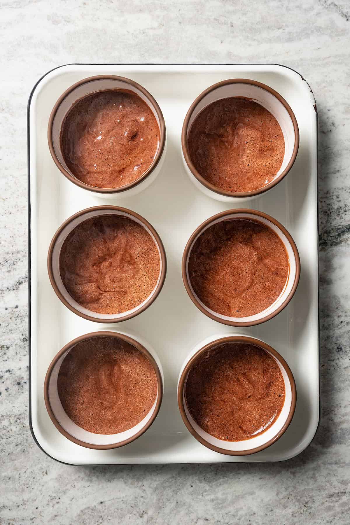 Six ramekins filled with chocolate souffle batter on a baking sheet.