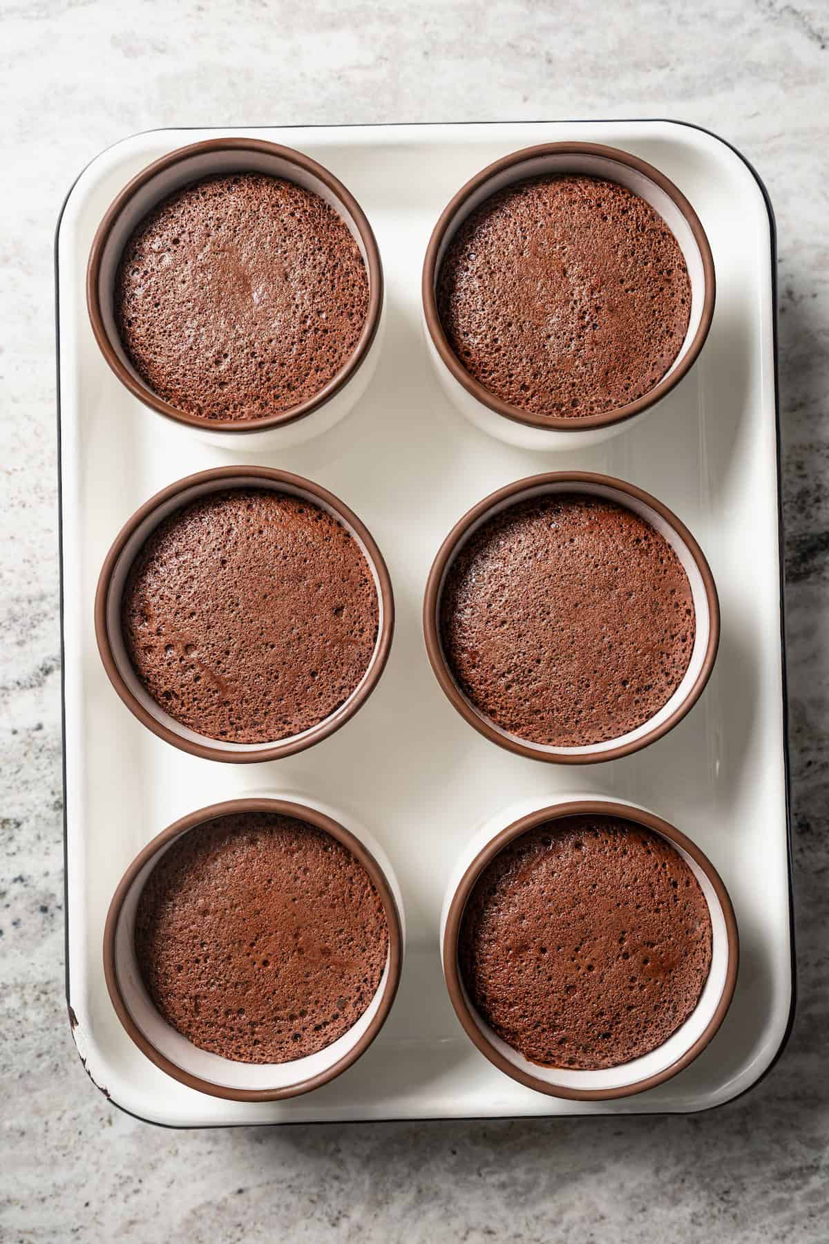 Six baked chocolate souffles in ramekins on a baking sheet.