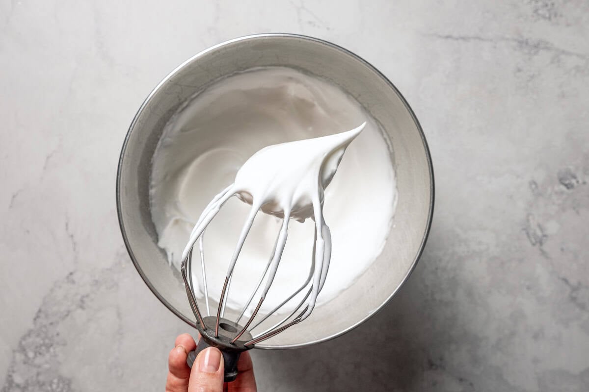 A hand holding a mixer's attachment with whipped egg whites over a bowl.