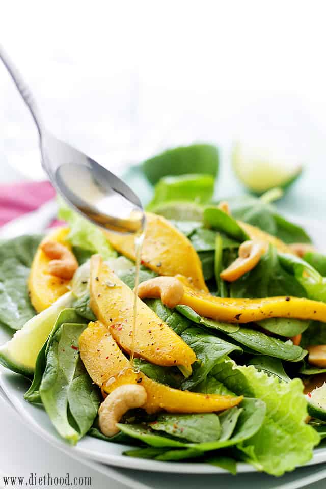 Pouring honey lime dressing over spinach leaves, mangoes, and cashews.