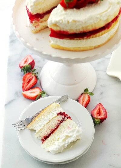 Overhead view of a slice of strawberry shortcake cake on a white plate, next to the rest of the cake on a white cake stand.