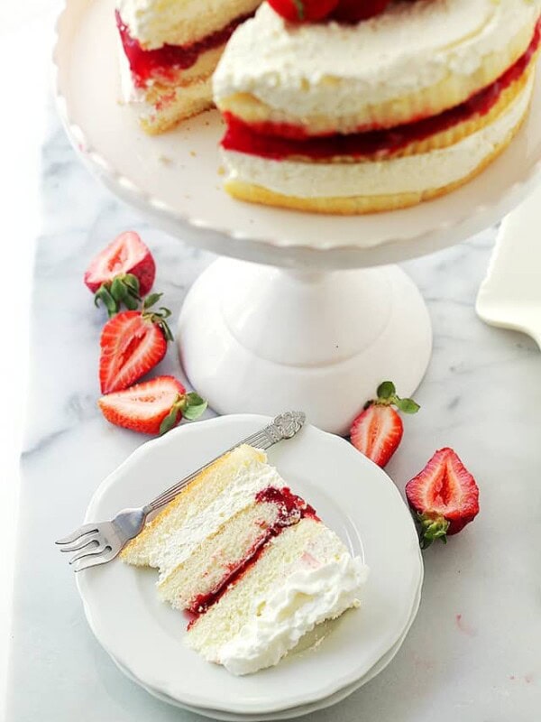 Overhead view of a slice of strawberry shortcake cake on a white plate, next to the rest of the cake on a white cake stand.