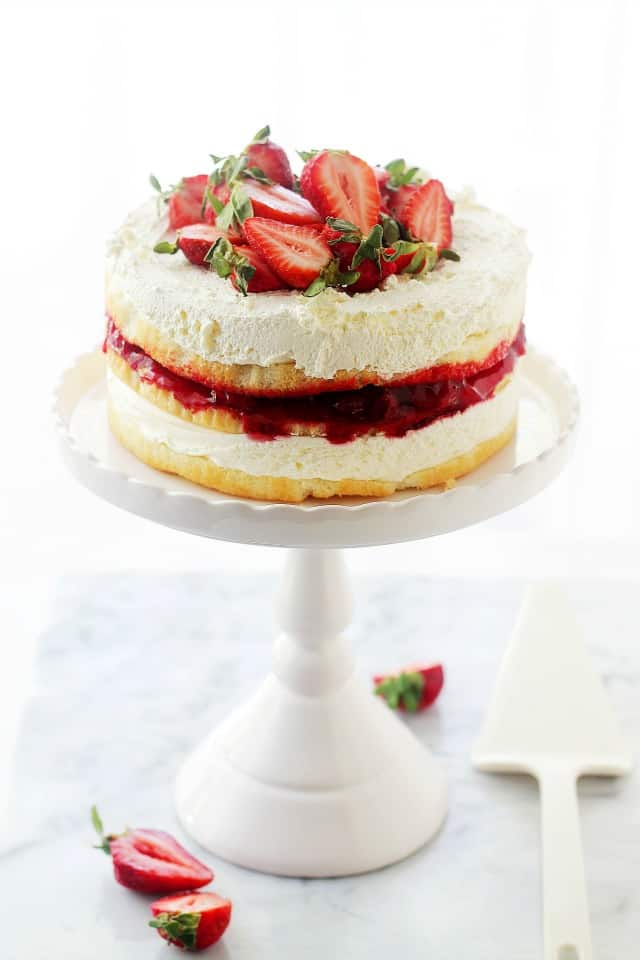 A strawberrry shortcake cake topped with whipped cream and fresh strawberries on a white cake stand.