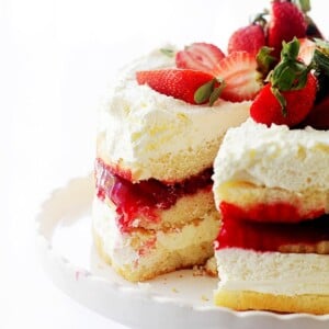 Strawberrry shortcake cake filled with strawberry filling and topped with whipped cream and fresh strawberries on a white cake stand, with a slice missing.