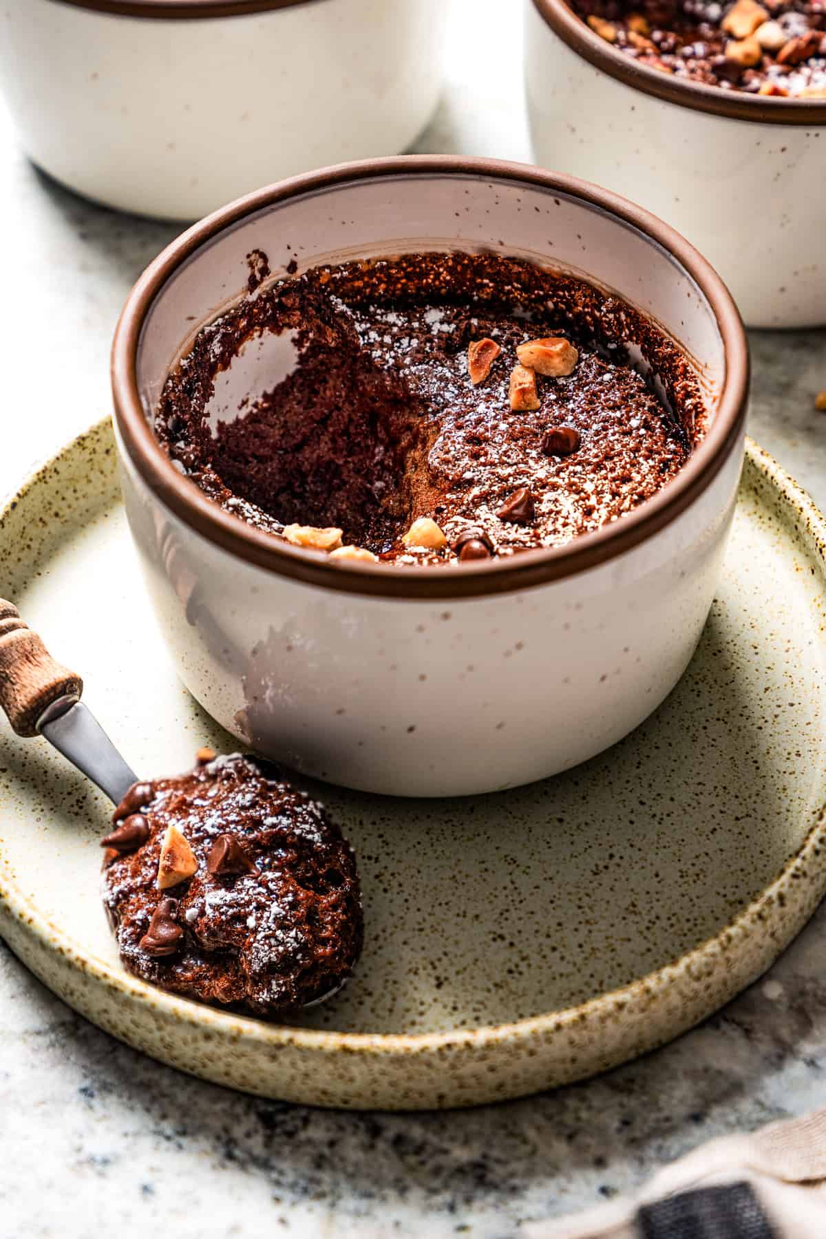 Chocolate souffle garnished with powdered sugar, chocolate chips, and hazelnuts in a ramekin with a spoonful missing.