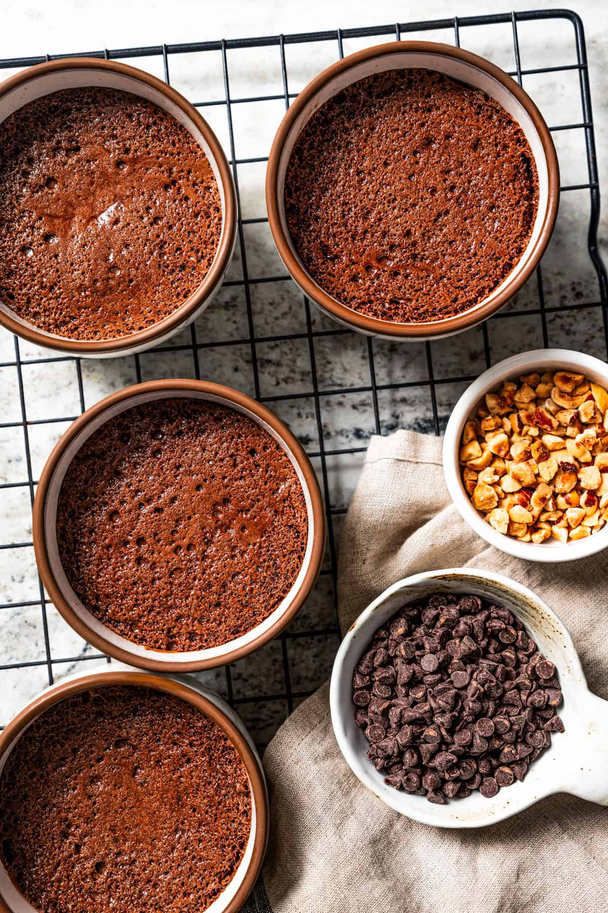 A wire rack with four chocolate souffles in ramekins, next to measuring scoops of chocolate chips and chopped hazelnuts.