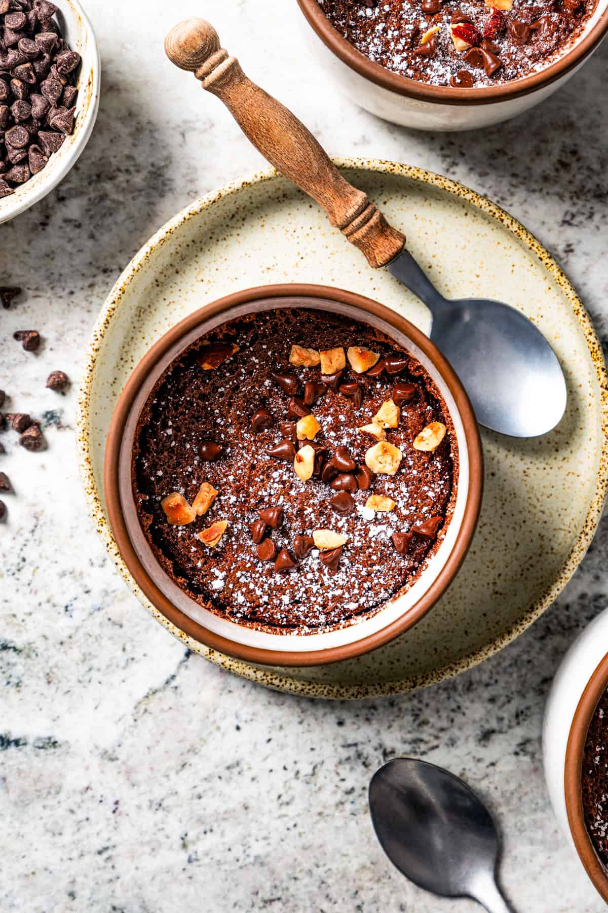 Chocolate souffle garnished with powdered sugar, chocolate chips, and hazelnuts in a ramekin on a plate beside a spoon.