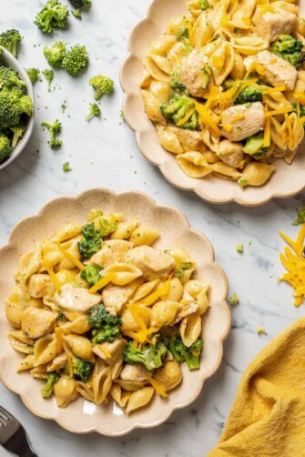 Overhead view of two plates of broccoli and chicken pasta, next to a bowl of broccoli, a fork, and a kitchen towel