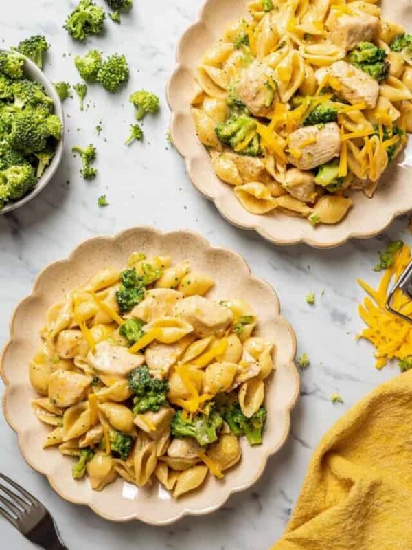 Overhead view of two plates of broccoli and chicken pasta, next to a bowl of broccoli, a fork, and a kitchen towel