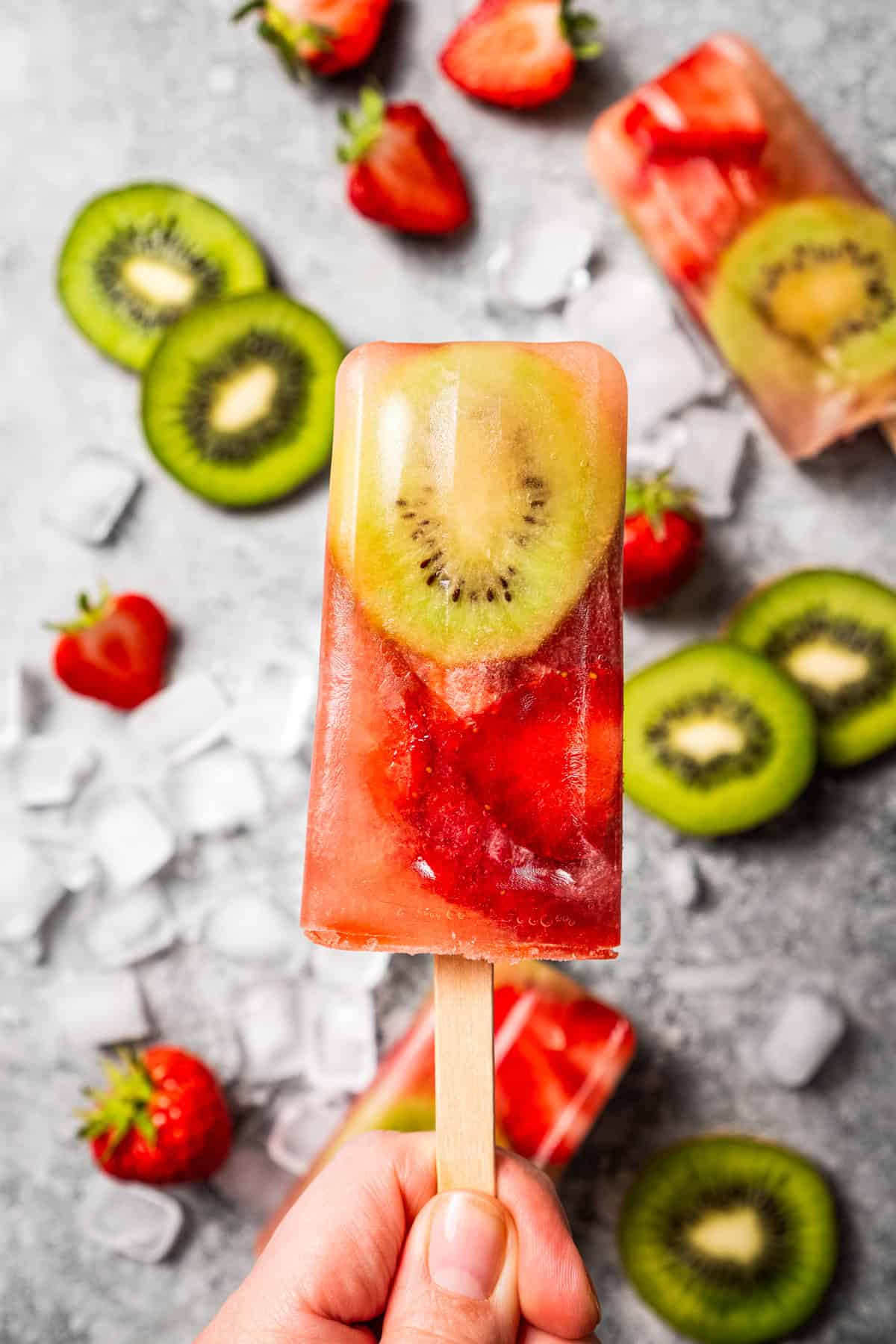 A hand holding a homemade fresh fruit popsicle, with sliced kiwis and strawberries in the background.