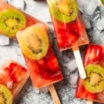 Assorted homemade fruit popsicles surrounded by sliced kiwis and strawberries.