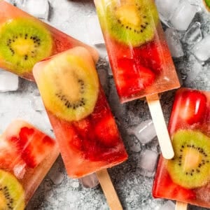 Assorted homemade fruit popsicles surrounded by sliced kiwis and strawberries.