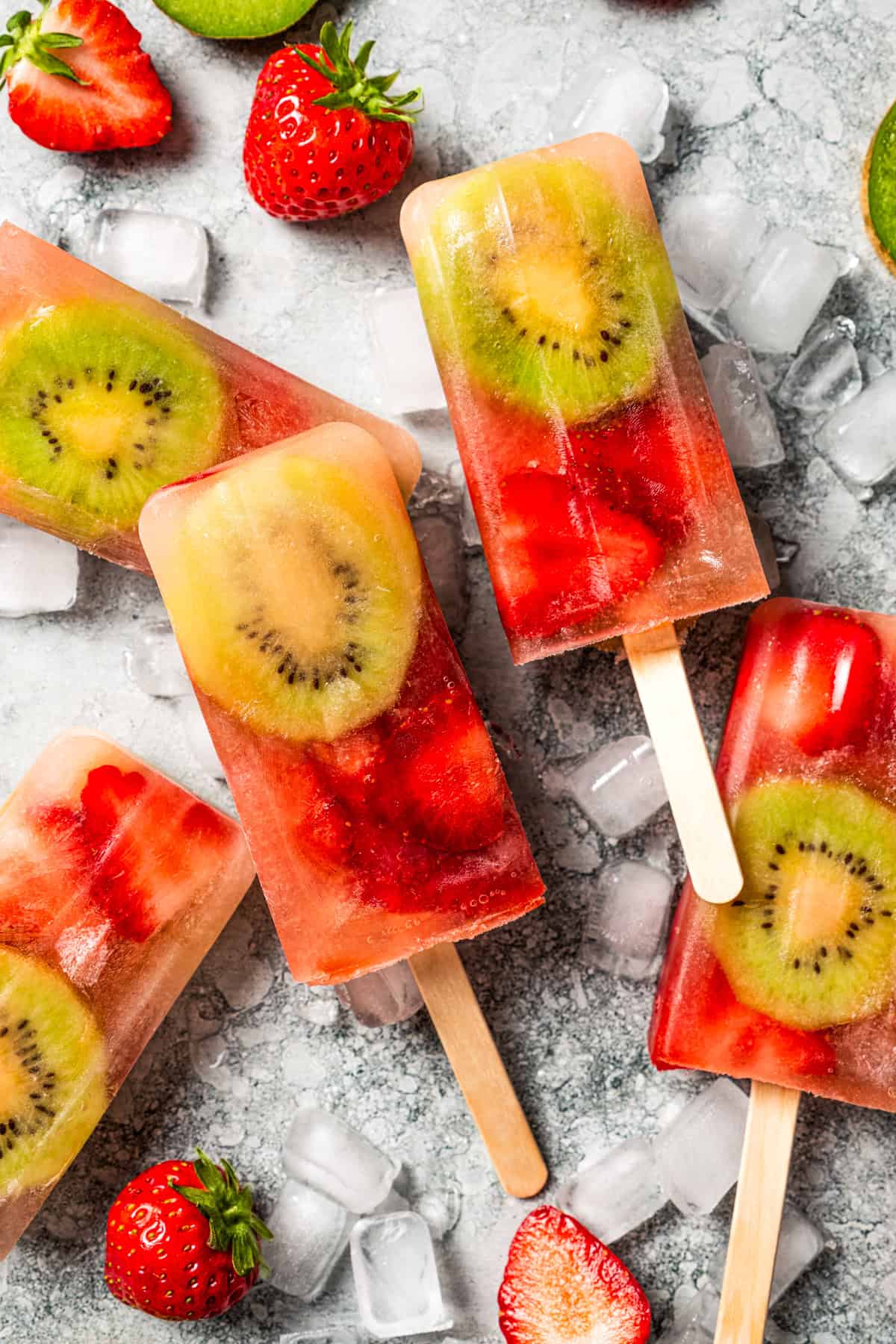 Assorted homemade fruit popsicles surrounded by sliced kiwis and strawberries.