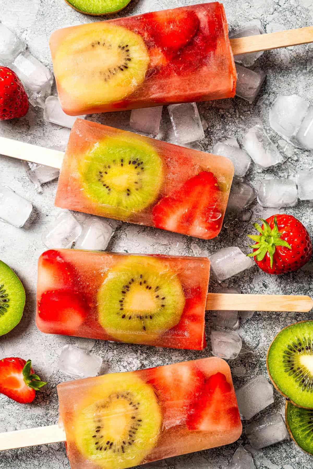 Four kiwi strawberry fruit popsicles surrounded by sliced kiwis and strawberries.