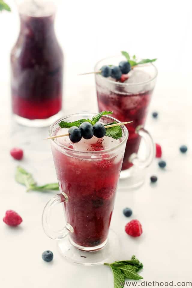 A tall glass of blueberry Agua Fresca with blueberries and mint on top and berries and mint on the table