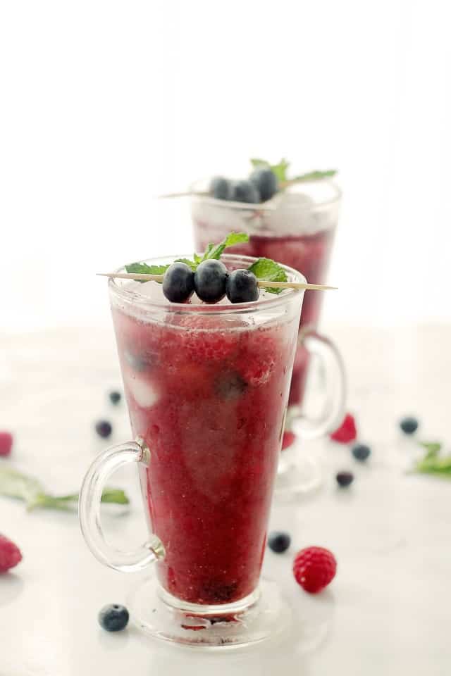 Side view of a glass of Agua Fresca with blueberries and mint on top and more berries and mint on the table