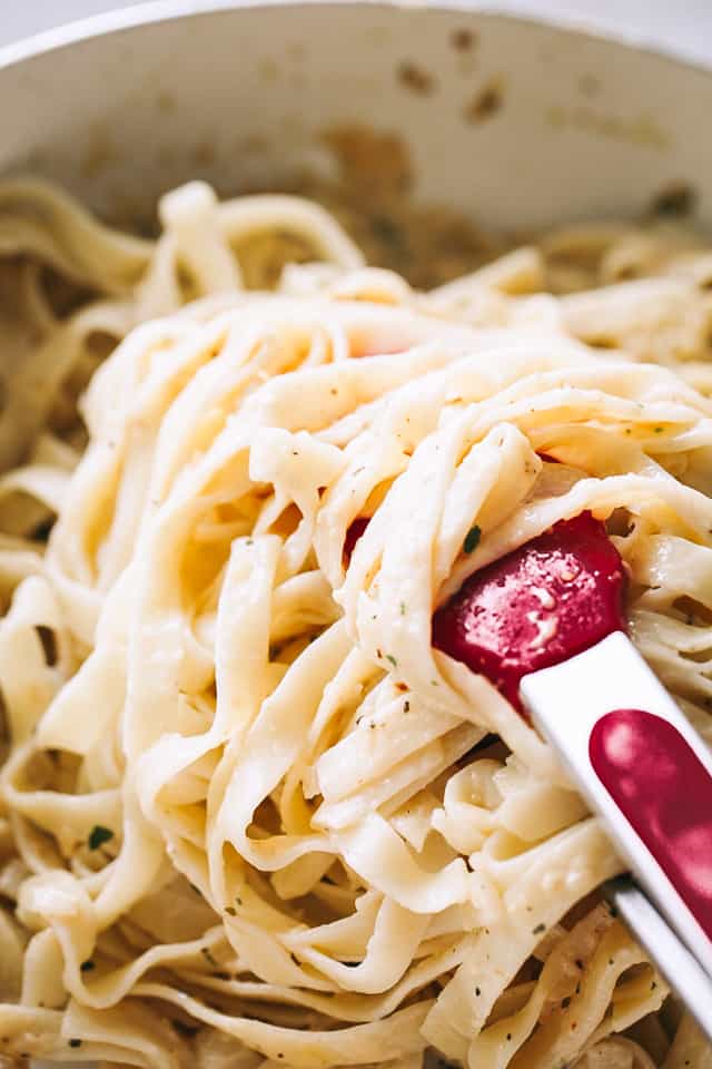 Cooked fettuccine pasta in a skillet with red tongs tossing the pasta with creamy alfredo sauce.