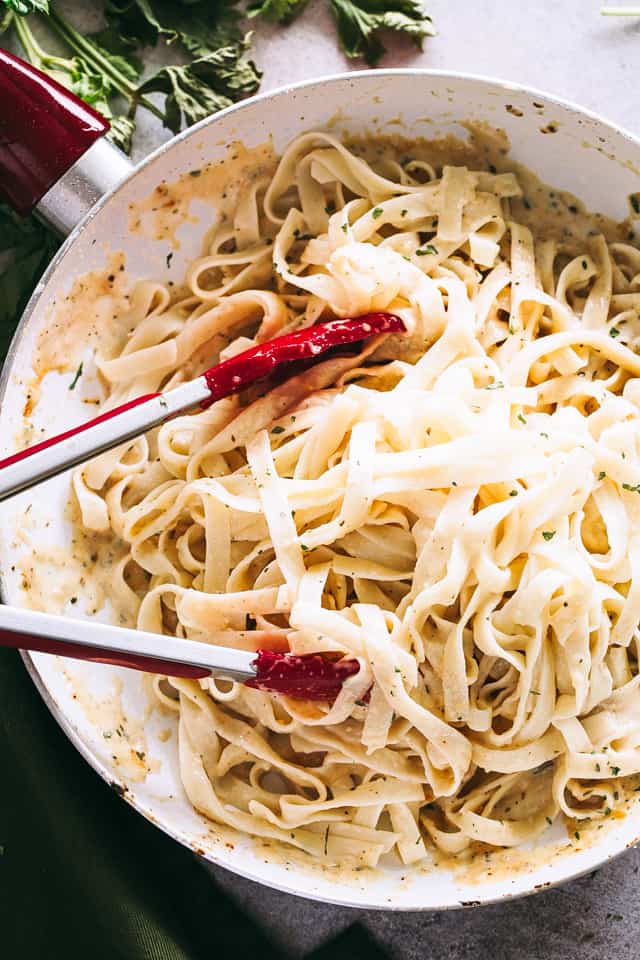 Cooked fettuccine pasta in a skillet with red tongs tossing the pasta with creamy alfredo sauce.