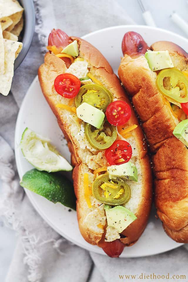 Hot Dogs in buns topped with sliced jalapenos, cheese, avocado, and tomatoes