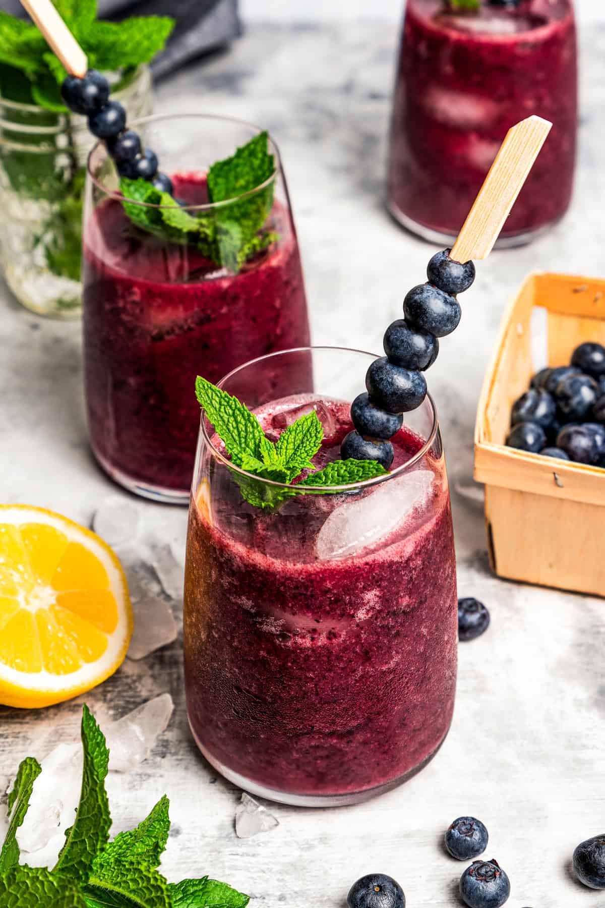A glass of agua fresca garnished with fresh mint and berries is set next to a tub of blueberries and half a lemon, with two more glasses in the background.