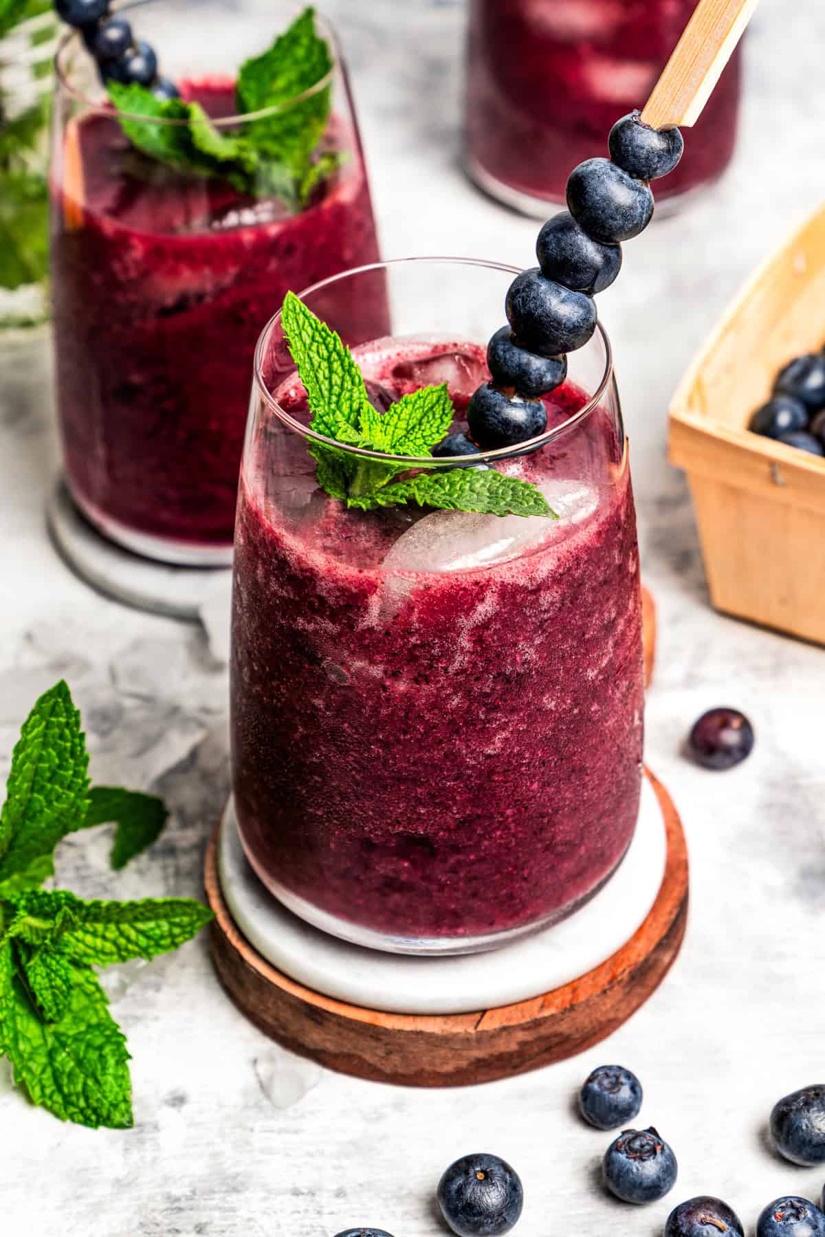 A glass of blueberry agua fresca garnished with fresh blueberries and mint is set on a wooden coaster, with two more glasses in the background.