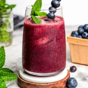 Blueberry agua fresca is served in a tall drinking glass set on a wooden coaster. It's garnished with fresh berries and mint.