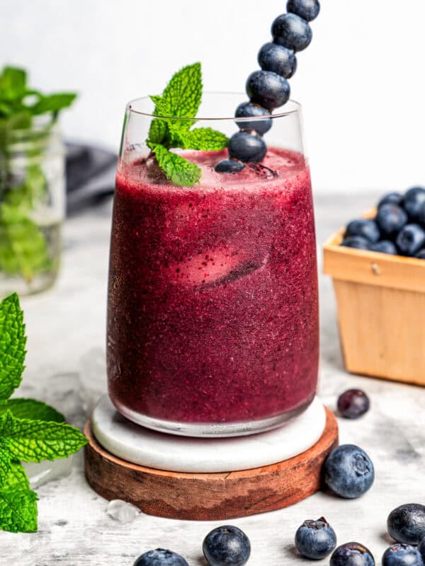 Blueberry agua fresca is served in a tall drinking glass set on a wooden coaster. It's garnished with fresh berries and mint.