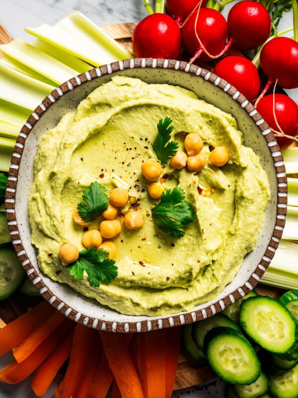 A bowl of hummus dip garnished with chickpeas, cucumber slices, and parsley, surrounded by crudites.