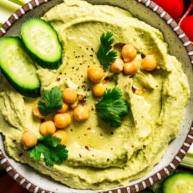A bowl of avocado hummus garnished with chickpeas, cucumber slices, and parsley, surrounded by crudites.