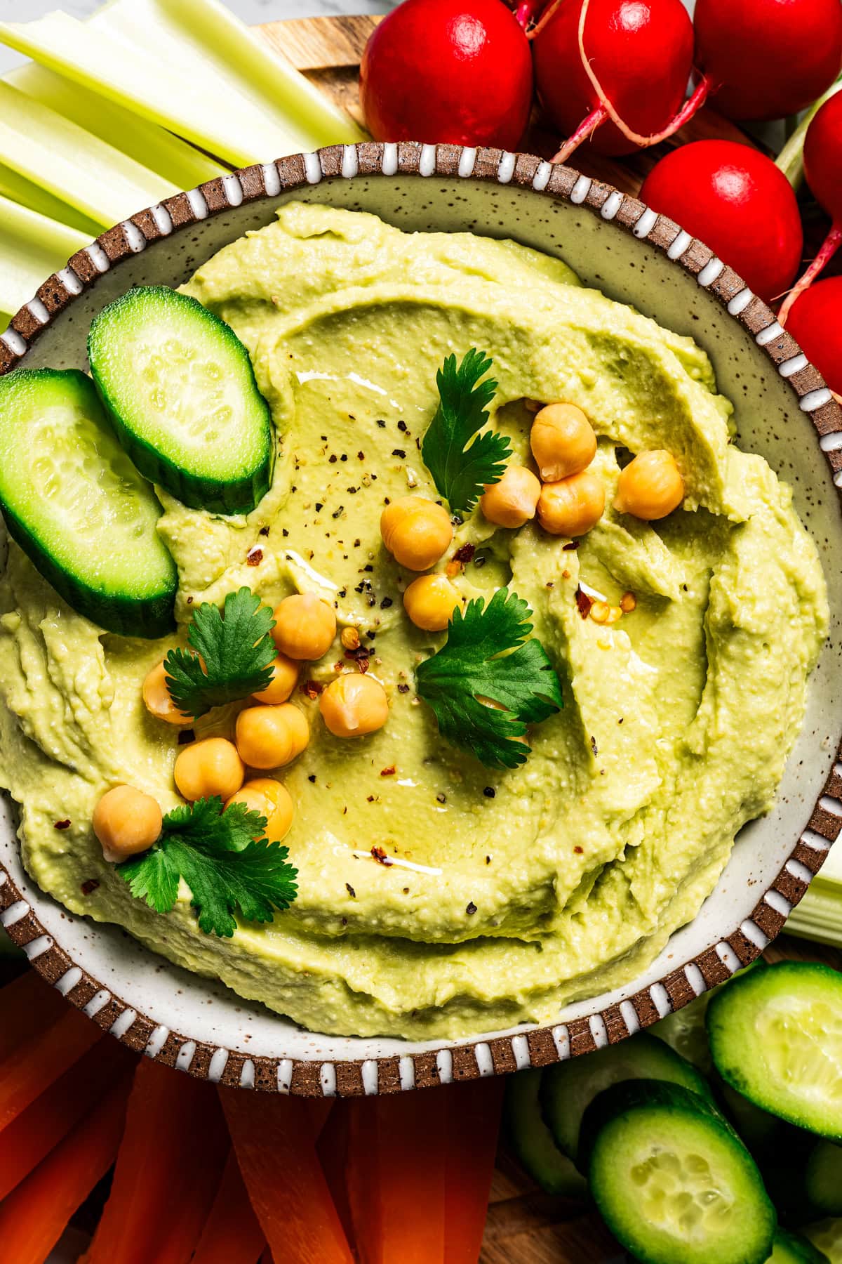 A bowl of avocado hummus garnished with chickpeas, cucumber slices, and parsley, surrounded by crudites.