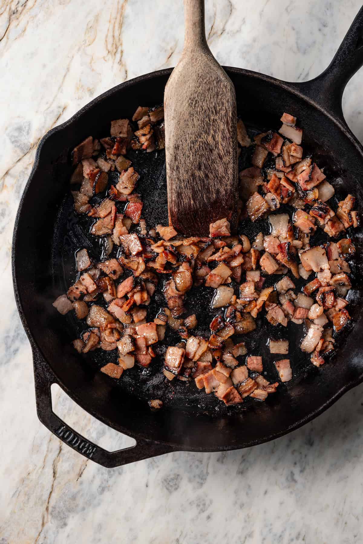 Cooked diced bacon being stirred in a skillet with a wooden spoon.