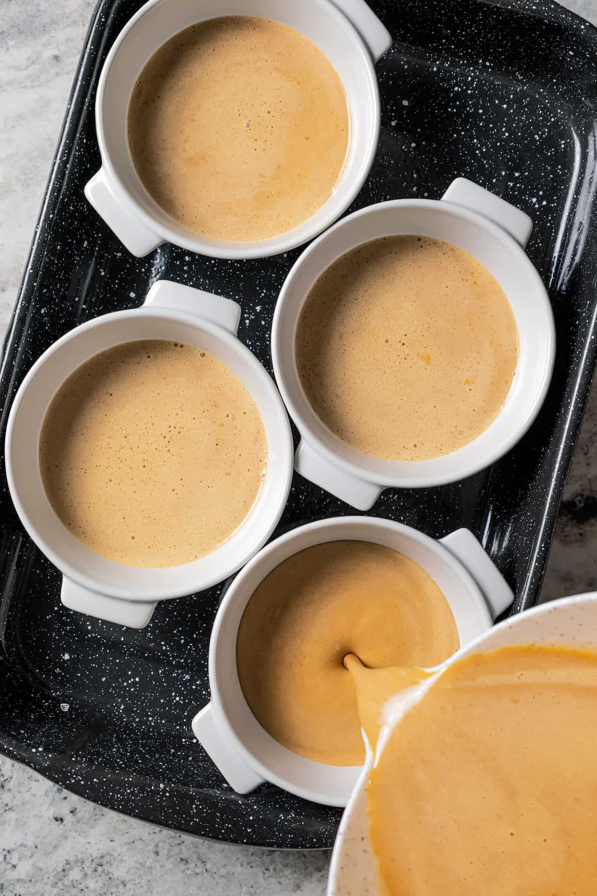 Coffee crème brûlée batter being poured into ramekins on a baking sheet.