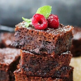 Three zucchini brownies stacked on a piece of parchment paper, garnished with fresh mint and raspberries, with more brownies in the background.