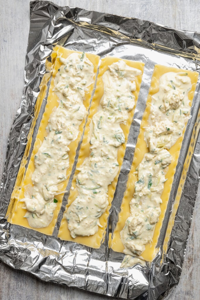 Three lasagna sheets topped with cheese and zucchini filling laid out on a foil-lined baking sheet.