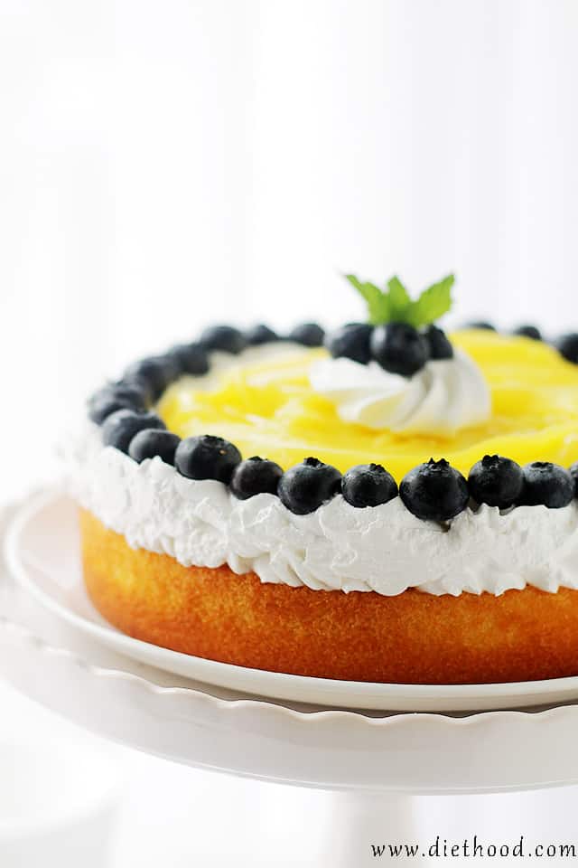 Side view of a lemon cake with lemon curd topped with whipped cream and blueberries on a white cake stand.