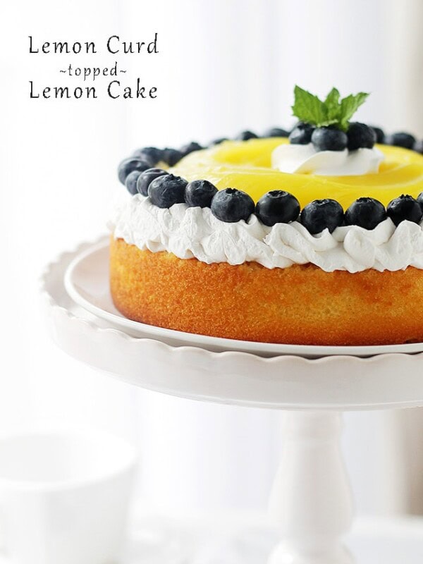 Side view of a lemon cake with lemon curd topped with whipped cream and blueberries on a white cake stand.