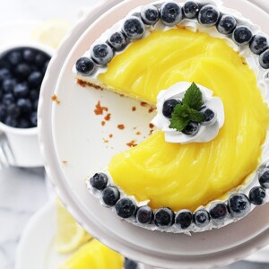 Overhead view of a lemon cake with lemon curd topped with whipped cream and blueberries on a white cake stand, with a slice missing.