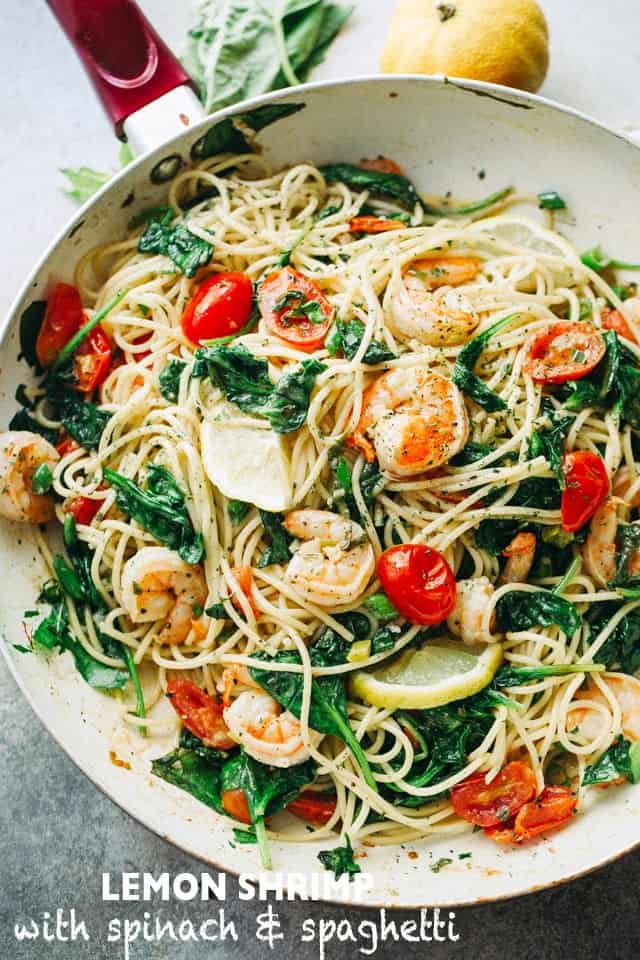 Overhead image of a skillet with cooked spaghetti, cherry tomatoes, baby spinach, shrimp, and thin slices of lemon.