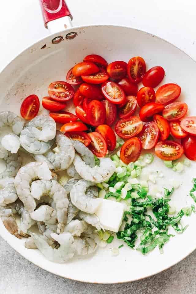 Raw shrimp, halved grape tomatoes, chopped green onions, and pats of butter in a white skillet.