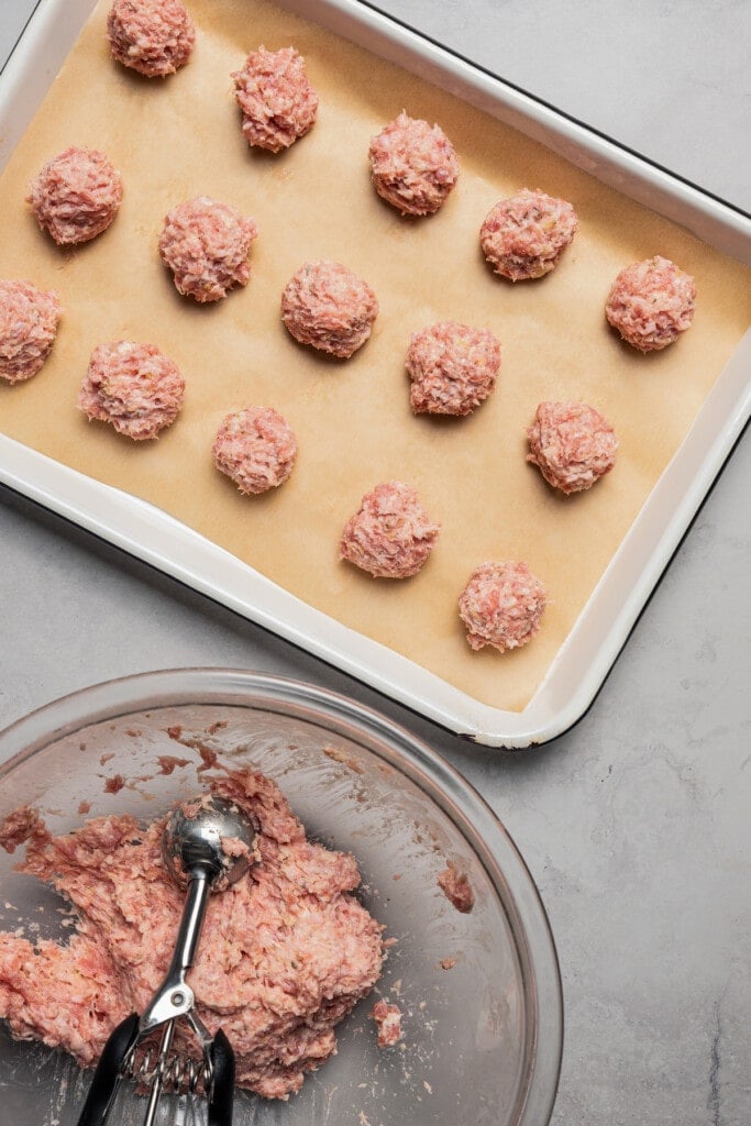 Rolled meatballs on a parchment-lined baking sheet next to a bowl with meat mixture.