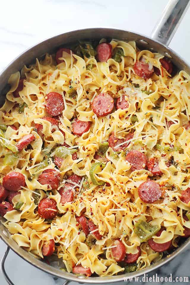 Overhead image of a skillet with cooked egg noodles, sliced turkey sausage, peppers, and tomatoes, and sprinkled with crushed red pepper flakes.
