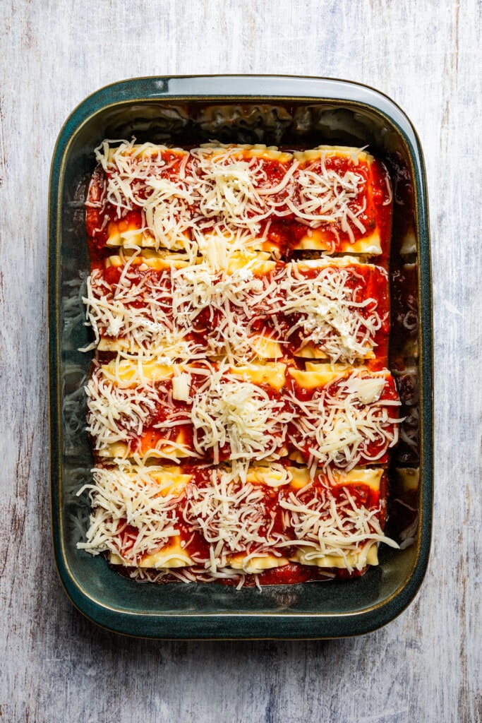 Assembled lasagna roll ups covered with tomato sauce and shredded cheese in a baking dish.