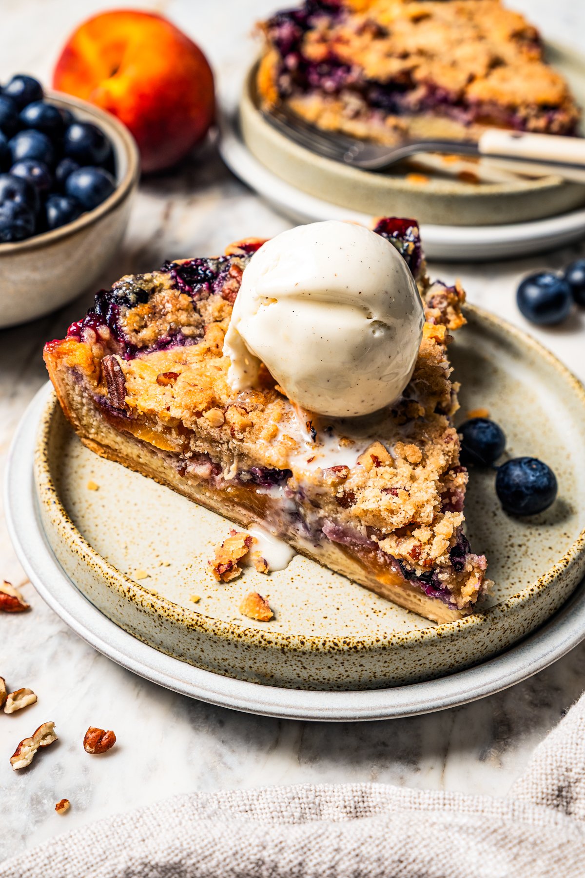 This peach blueberry pie is a fruit-filled custard pie bursting with juicy fresh peaches and blueberries, topped with a nutty pecan streusel. A slice of peach blueberry pie topped with a scoop of vanilla ice cream on a plate.