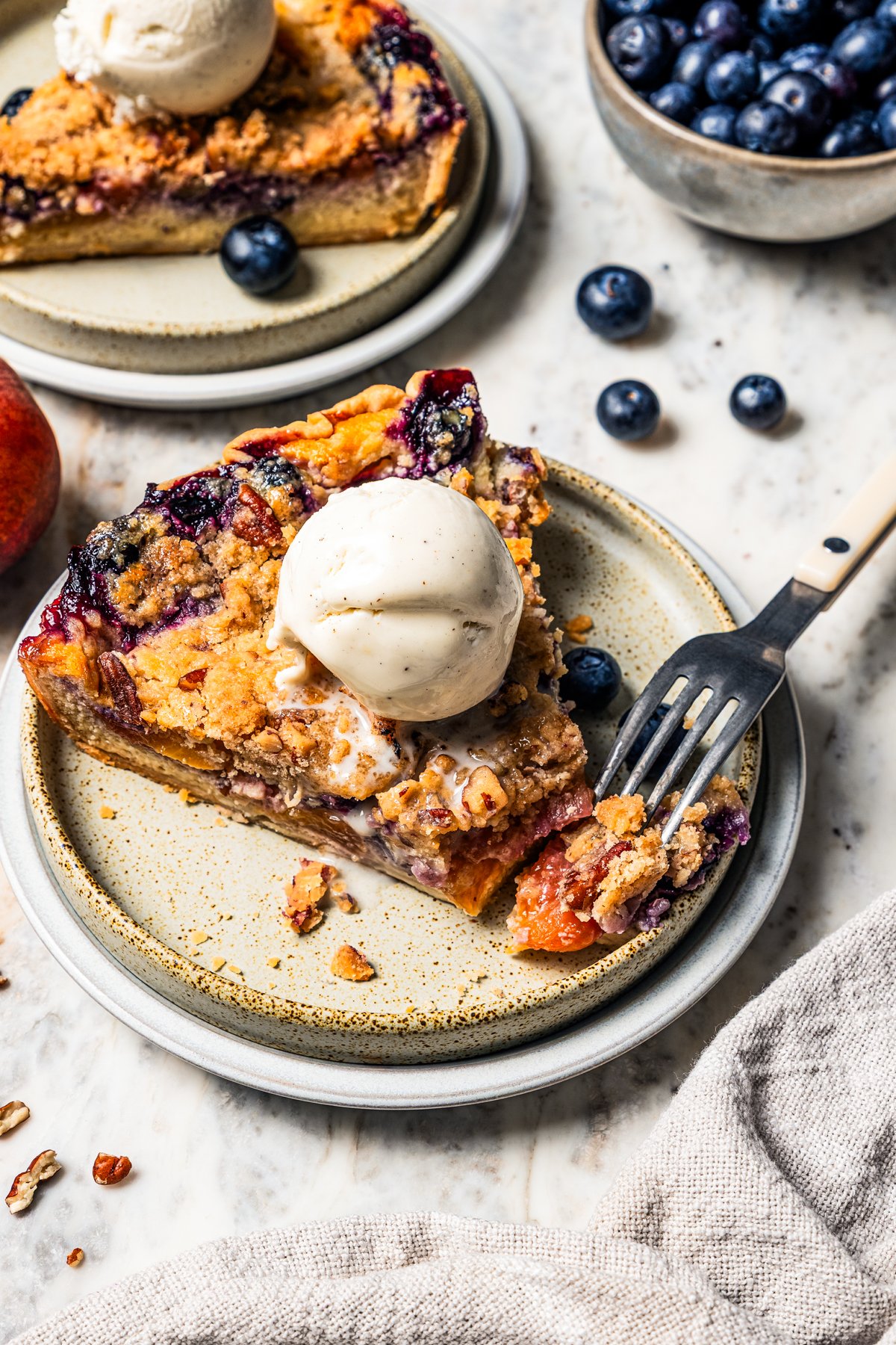 This peach blueberry pie is a fruit-filled custard pie bursting with juicy fresh peaches and blueberries, topped with a nutty pecan streusel. A fork resting on the end of a slice of peach blueberry pie, topped with a scoop of vanilla ice cream on a plate. photo by diethood.com