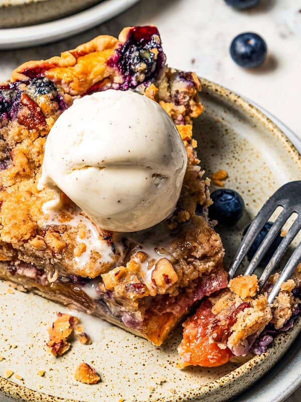 A fork resting on the end of a slice of peach blueberry pie, topped with a scoop of vanilla ice cream on a plate. photo by diethood.com